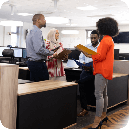 Multiple people in a government office consulting to one another around desks.