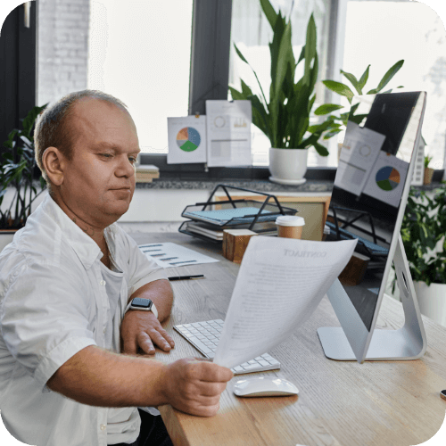 person with disability at a desk in front of a large monitor screen holding a white piece of paper